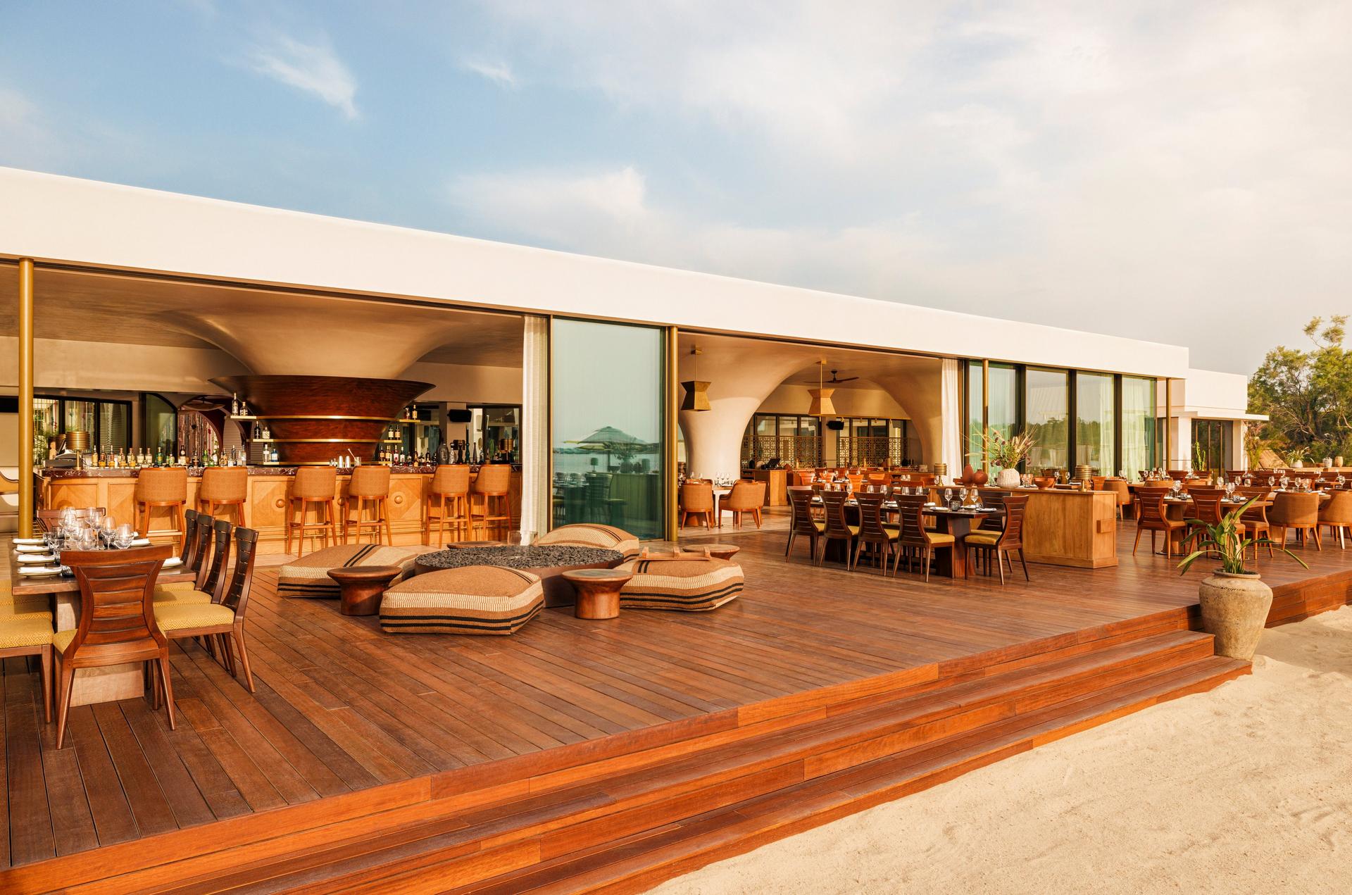 The open terrace at Manko, One&Only Aesthesis: a teak deck with low fire tables, the bar behind, sand at the edge of the frame.